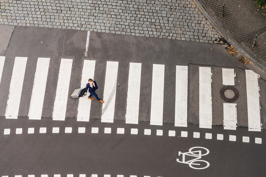 Germany, businesswoman with rolling suitcase walking on zebra crossing, view from above