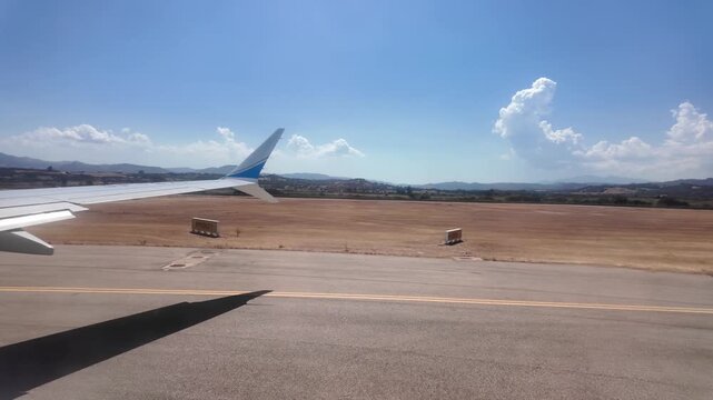 Passenger aircraft taxiing on an airport runway on a sunny day; a view from the cabin window showing the wing with a winglet and yellow navigation markings.