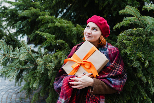 Dreaming woman holds a wrapped holiday gift near a winter fir tree