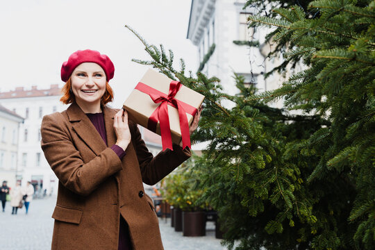 Cheerful woman holds a wrapped gift by a Christmas tree outdoors