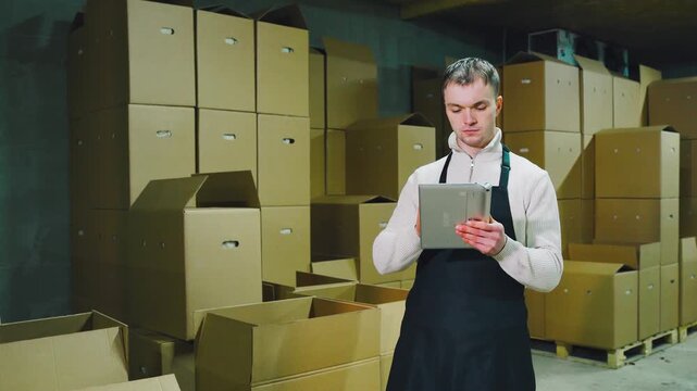 Focused warehouse worker in dark apron checks inventory with tablet beside stacked cardboard boxes on pallets inside dim storage room. Farmer grower dispatches products for sale. Small business.