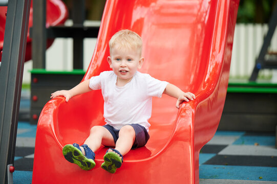 Child sliding down playground slide, holding sides and smiling, summer outfit, bright red slide in park, outdoor activity, fun childhood, active play