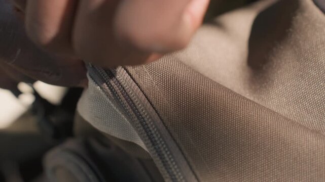 Close up of hand interacting with zipper on outdoor backpack. fabric shows detailed texture and stitching indicating durable material. Soft lighting highlights interaction between human skin