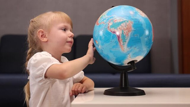 Little blonde girl spinning globe model and exploring world map at home