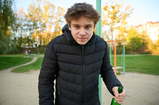 Teenage male stands outdoors holding bottle in hand. Urban park background and hostile expression suggest emotional conflict and unhealthy choices. Concept of youth addiction warning