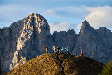 Group of hikers gathers on narrow sunlit ridge beneath towering Dolomite cliffs in Italy,...