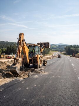 A backhoe loader dumps material while digging alongside a newly paved road, with workers and rolling countryside visible in the background.
