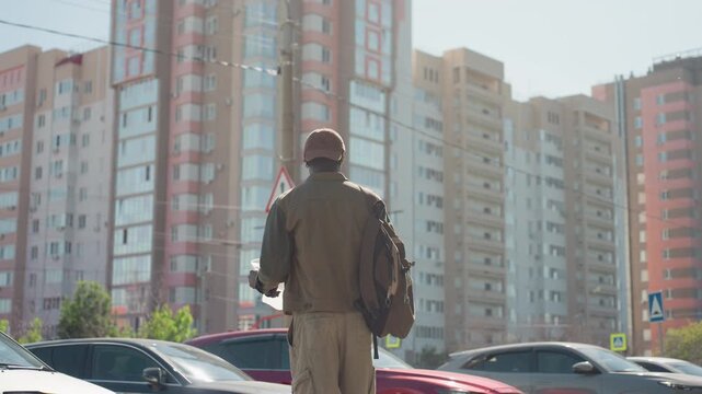 Man holding map and backpack walks through urban street with parked cars tall apartment buildings in background. Man examines paper while moving along sidewalk under clear daylight wearing