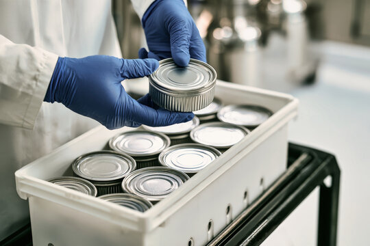 A worker in blue gloves and a white coat places a ribbed silver can into a white plastic container in a factory environment. Six cans are lined up