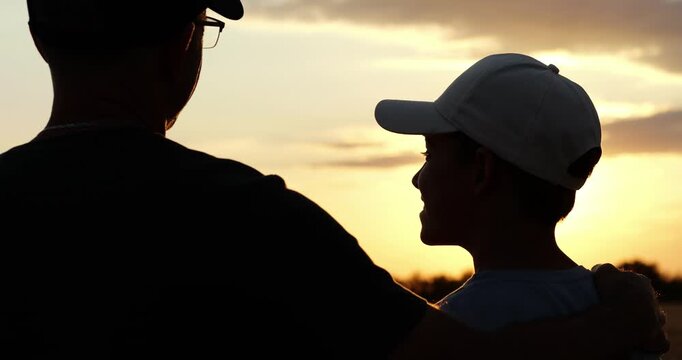 Father and son watch sunset in silhouette. Parent and child share quiet outdoor moment at golden hour. Loving family bond evokes connection, hope, and childhood memories in rural landscape.