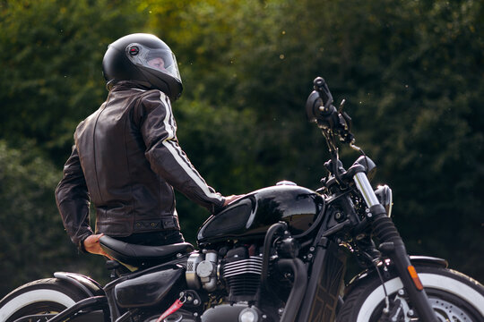 Motorcyclist wearing a black helmet and leather jacket, standing next to a classic black motorcycle with a blurred green background