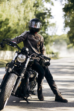 Biker wearing leather jacket and helmet, resting on a classic motorcycle parked on a quiet road, enjoying freedom and travel