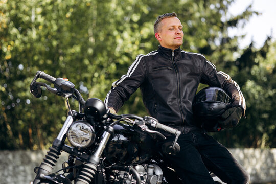 Male motorcyclist wearing leather jacket and gloves, holding a black helmet while sitting on his bike, looking away