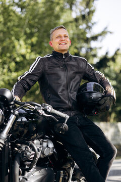 Man in leather jacket and jeans sitting on a custom black motorcycle, holding a helmet and smiling outdoors