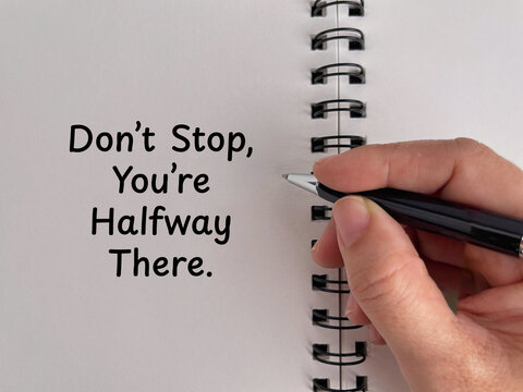 Hand writing &ldquo;Don&rsquo;t Stop, You&rsquo;re Halfway There&rdquo; in a notebook, representing motivation, perseverance, progress, determination, and goal achievement.