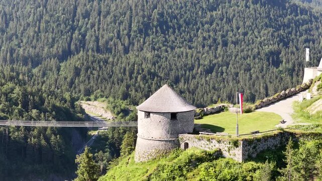 Ehrenberg castle watchtower stands above forested valley near Reutte, Austria, aerial drone orbit around stone turret, flag and suspension bridge.