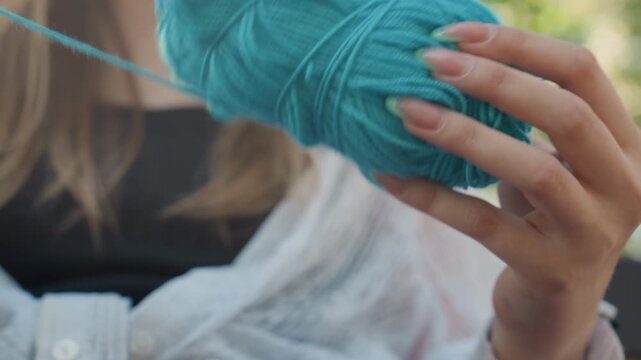 Close up of young woman untangling turquoise yarn with long nails revealing texture detail and soft lighting. scene focuses on hands manipulating thick blue thread against outdoor blurred