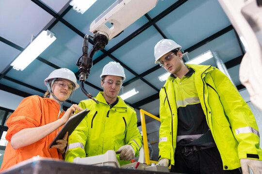 Industrial engineering team wearing safety jackets and hard hats analyzing robotic arm system in automated factory using tablet and control panel for quality control, smart manufacturing process.