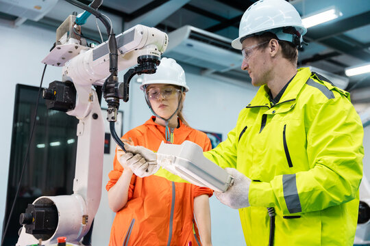 Male engineer in hardhat and safety glasses uses teach pendant to control an industrial robot arm female technician observes, showing automation training, factory safety and robotics.