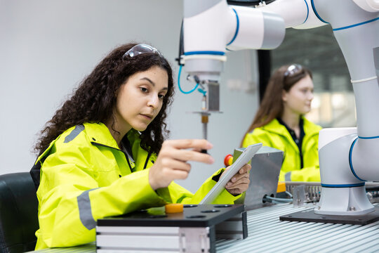 Female engineer in safety jacket uses a tablet and control buttons beside a collaborative robot arm, showing robotics programming, automation training, smart manufacturing, and quality control.