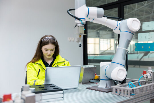 Female engineer in safety glasses works on laptop beside collaborative robot arm, showing robotics programming, industrial automation, smart manufacturing, quality control.