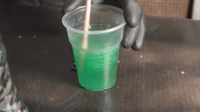 A person in black glove stirs liquid green epoxy resin with glitter filler using a wooden stick in a plastic cup, close-up shot.