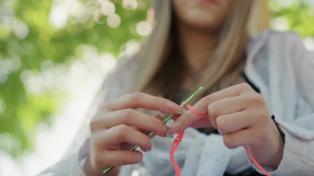Close up of woman crocheting with pink yarn and green hook hands in focus against blurred outdoor background. Soft natural light highlights texture of thread as woman works on textile craft