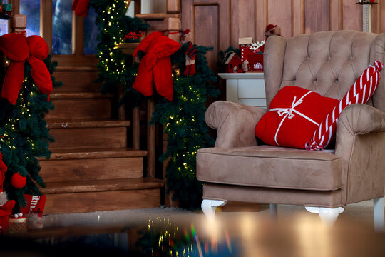 Warm ambient lighting illuminates a comfortable armchair with a red gift pillow beside a wooden staircase adorned with evergreen garlands and bows