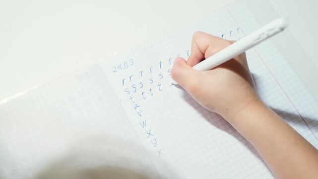 Child practices writing English alphabet letters by hand on graph paper with pen, building early literacy and handwriting skills through focused learning exercise and repeated letter formation