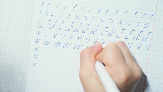 Child practices writing English alphabet letters by hand, tracing rows of lowercase forms with blue pen on squared notebook paper during early handwriting lesson, repeating strokes to learn letter