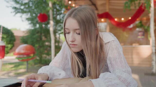Close up of young woman writing with pen on notebook at outdoor table. Woman wears white lace top and black inner shirt long blonde hair visible. Background features green trees red string