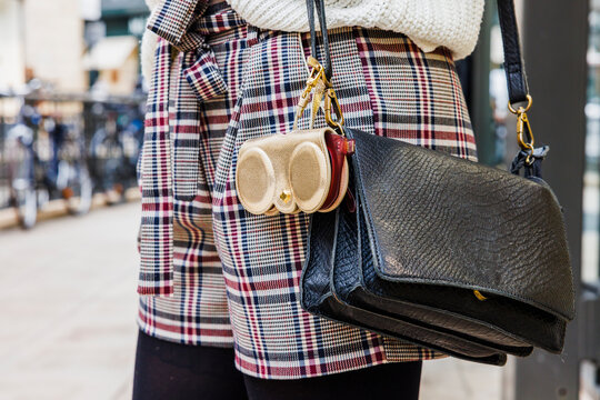 D&eacute;tail d&rsquo;un sac &agrave; main noir port&eacute; avec une veste &agrave; carreaux, accompagn&eacute; d&rsquo;un petit accessoire dor&eacute; fix&eacute; &agrave; la bandouli&egrave;re.