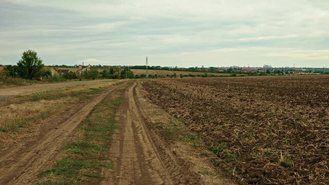 A wide shot shows a rural dirt road running alongside a plowed agricultural field under a cloudy overcast sky.