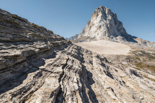 Layered sedimentary rock formation with smooth flat and deeply grooved surfaces in light beige to gray, a broad pale barren expanse beyond and a towering striated peak in the distance