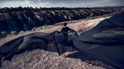 Obraz premium Fearless figure extends black fabric like wings beneath dramatic sky with commanding energy. Personal power, freedom, cinematic fantasy concept