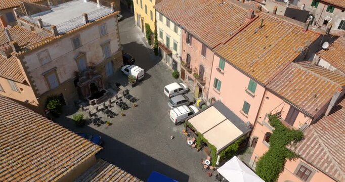 Aerial view of a small square in the historic center of Tuscania, in the province of Viterbo, in Lazio, Italy, with outdoor cafe tables, an awning, parked vans and pink and yellow facades with ivy.