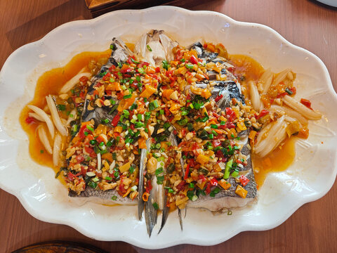 dish of steamed bighead carp fish head smothered in a rich, savory, and fiery sauce of chopped pickled red and green chilies, minced garlic, and sliced ginger on a white platter