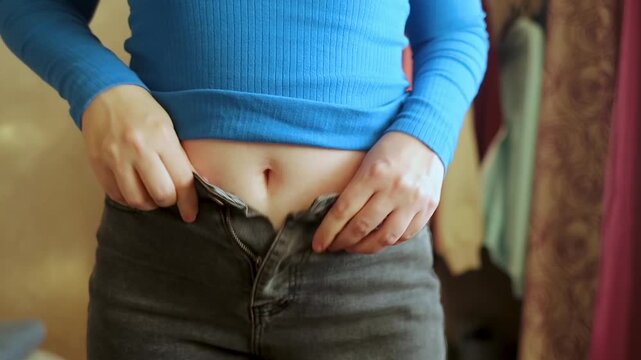 Overweight woman struggling to button tight jeans after postpartum body changes. Concept of weight loss, belly fat, and post pregnancy body positivity.