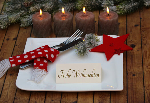 Festlicher Weihnachtstisch mit vier brennenden Adventskerzen, Teller und Besteck auf rustikalem Holztisch und einer Karte mit der Aufschrift Frohe Weihnachten.
