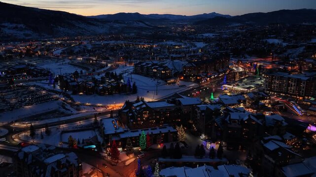 Winter drone shot over a ski resort town resembling Aspen, Colorado, shows snow covered rooftops, glowing roads, festive holiday lights, and surrounding mountain terrain after fresh snow.