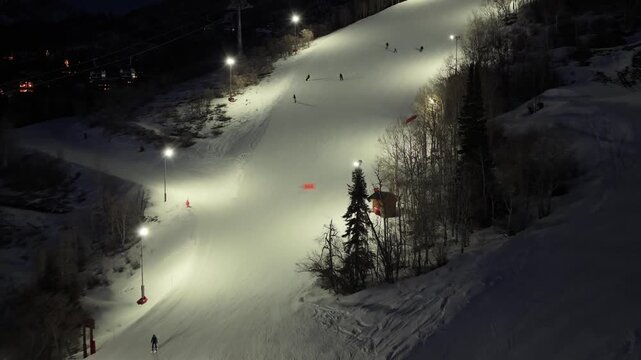 After-dark aerial of Steamboat Springs, Colorado shows night skiers moving across glowing slopes, gondola lifts over the snow, forested terrain, and floodlights illuminating the ski runs.