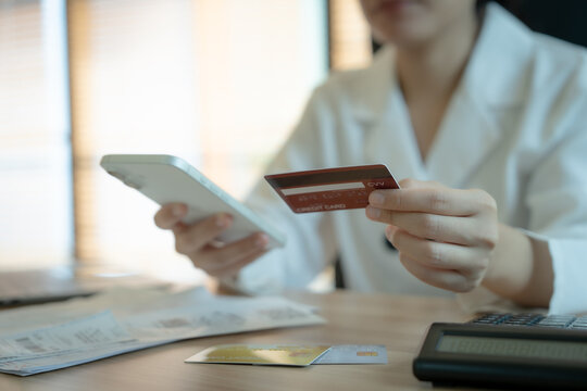 Woman holds card and phone at desk. She appears focused and calm. Documents, calculator, and credit card lie nearby. She likely manages finances or makes online purchases