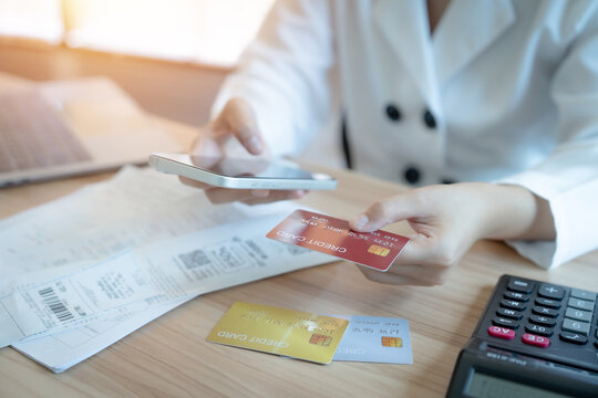 Person holds phone and credit card near documents. Hands gesture toward calculator and receipts. Warm light bathes wooden desk surface. Focus on payment or financial management
