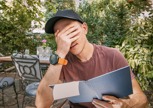 Man covering face in shock and frustration after receiving a surprisingly expensive restaurant bill while dining outdoors