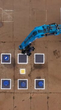 A top-down aerial view shows a large blue material handler hydraulic crane arm swinging over an asphalt training or testing area marked with squares and barrels.