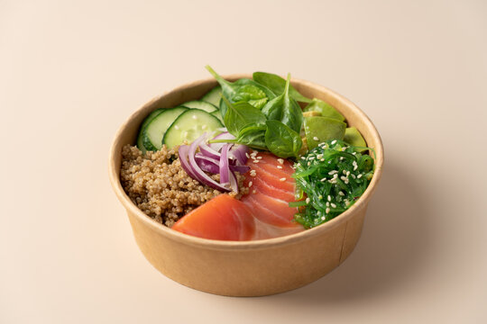 Healthy protein bowl with quinoa, smoked salmon, spinach, cucumber, avocado and chuka seaweed salad in a cardboard container on a light beige background
