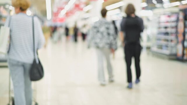 Individuals are walking in an indoor shopping area filled with shelves. The setting shows a blurred perspective of people in casual clothing moving through the grocery aisle of a shopping mall.
