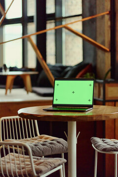 Vertical shot of open laptop with green screen mockup standing on table in coworking cafe. Modern device ready for digital website presentation, software interface advertising