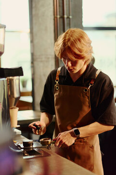 Vertical shot of young Caucasian male barista tamping ground coffee in portafilter at cafe counter. Image suitable for coffee shop marketing and small business advertising