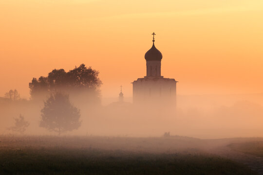 Silhouette of ancient Russian Orthodox church above misty field backlit by rising sun, Russia